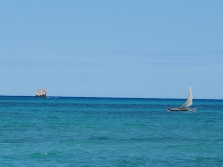 A small sailboat with a white sail travels across the turquoise ocean. In the background, a rocky islet is visible under a clear blue sky.