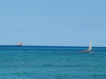 A small sailboat with a white sail travels across the turquoise ocean. In the background, a rocky islet is visible under a clear blue sky.
