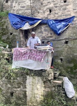 An older man is standing at a makeshift outdoor ironing station set against a weathered stone wall with patches of greenery and moss. A blue tarp provides partial shelter overhead, while a covered ironing board sits in front of him. A younger person is seen sitting nearby. The scene suggests a humble and improvised setup for laundry work.
