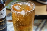 A close-up view of a glass filled with a fizzy amber beverage with ice cubes, set on a table next to a partially visible bottle. The effervescence and transparency of the drink are accentuated by the light forming bubbles and reflections.