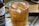 A close-up view of a glass filled with a fizzy amber beverage with ice cubes, set on a table next to a partially visible bottle. The effervescence and transparency of the drink are accentuated by the light forming bubbles and reflections.