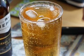 A close-up view of a glass filled with a fizzy amber beverage with ice cubes, set on a table next to a partially visible bottle. The effervescence and transparency of the drink are accentuated by the light forming bubbles and reflections.