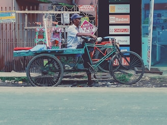 A person is sitting on a bicycle rickshaw parked on the side of the road. The rickshaw is painted in bright colors, and there are small decorative items displayed behind the seat. Advertisements for electronics brands such as Realme, Redmi, Oneplus, and iPhone are visible in the background, along with signs indicating a no parking zone.