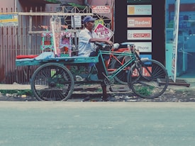 A person is sitting on a bicycle rickshaw parked on the side of the road. The rickshaw is painted in bright colors, and there are small decorative items displayed behind the seat. Advertisements for electronics brands such as Realme, Redmi, Oneplus, and iPhone are visible in the background, along with signs indicating a no parking zone.