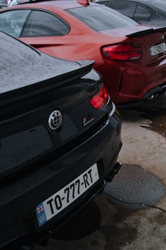 Two parked cars with different color schemes, one black and one red, possibly BMW models, are seen from their rear ends. Both cars have visible license plates, with the black car showing 'TO-777-RT' and the red car showing 'MZ-2022-01'. The ground has patches of water, indicating recent rain, and the cars appear to have water droplets on their surfaces.