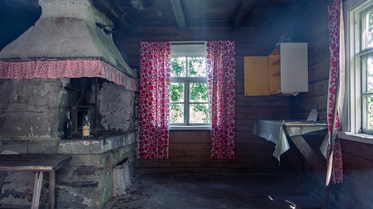 A rustic room features a large, old fireplace with bottles placed on it. Red patterned curtains hang at the window, filtering in natural light. The wooden walls and ceiling give the space a cozy, vintage feel. A simple, small table and old cabinet complete the setting.