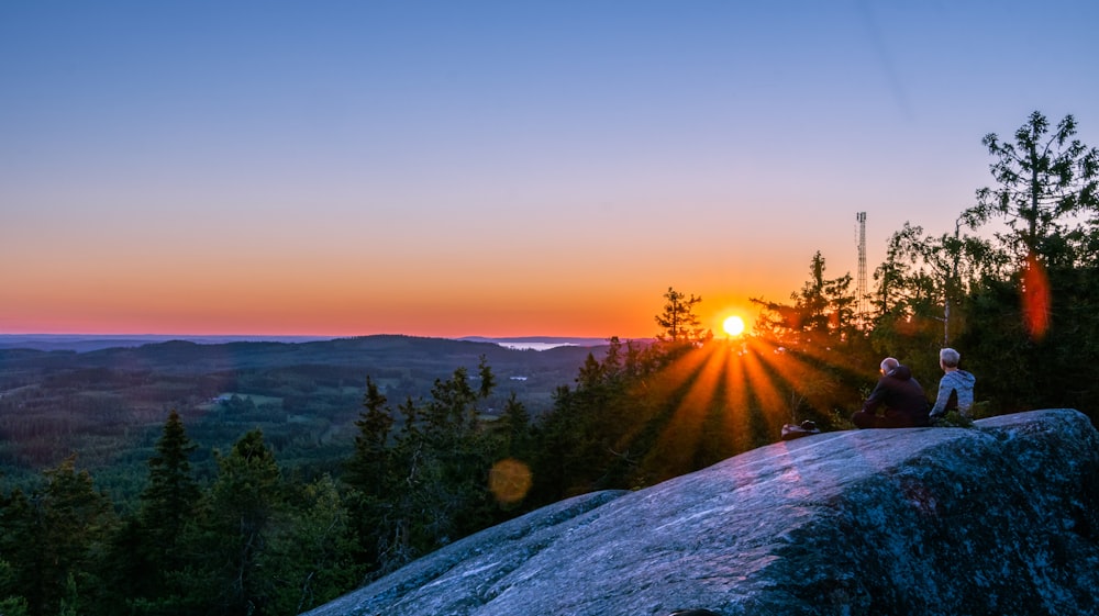 a couple people sitting on a rock overlooking a forest and sunset