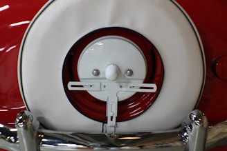 A close-up view of a vintage car's rear featuring a white and red spare tire cover and chrome bumper. The white circular cover is set against a red background, with shiny chrome accents visible at the bottom.