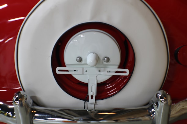 A close-up view of a vintage car's rear featuring a white and red spare tire cover and chrome bumper. The white circular cover is set against a red background, with shiny chrome accents visible at the bottom.
