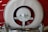 A close-up view of a vintage car's rear featuring a white and red spare tire cover and chrome bumper. The white circular cover is set against a red background, with shiny chrome accents visible at the bottom.
