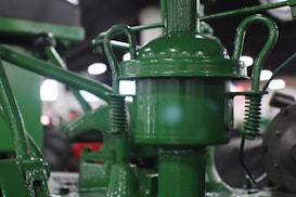 A close-up view of an old mechanical device with various metal components. The device is painted green and features a cylindrical central part with springs and rods attached to it. The background is blurred, with lights suggesting an indoor setting.