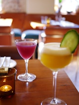 Two elegantly presented cocktails stand on a wooden table in a cozy restaurant setting. The glass in the foreground contains a yellow drink garnished with a slice of cucumber, while the other glass holds a vibrant pink beverage. A small candle in a brown glass holder adds a warm ambiance to the scene.