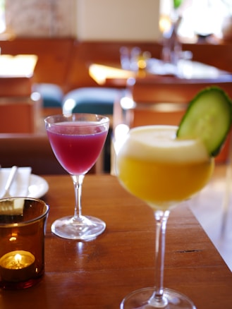 Two elegantly presented cocktails stand on a wooden table in a cozy restaurant setting. The glass in the foreground contains a yellow drink garnished with a slice of cucumber, while the other glass holds a vibrant pink beverage. A small candle in a brown glass holder adds a warm ambiance to the scene.