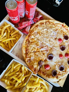 A meal spread featuring a large pizza topped with cheese, onions, and various meats. Surrounding the pizza are several boxes of golden, crispy French fries. Three cans of Coca-Cola Classic are placed nearby, along with a stack of ketchup sachets.