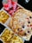 A meal spread featuring a large pizza topped with cheese, onions, and various meats. Surrounding the pizza are several boxes of golden, crispy French fries. Three cans of Coca-Cola Classic are placed nearby, along with a stack of ketchup sachets.