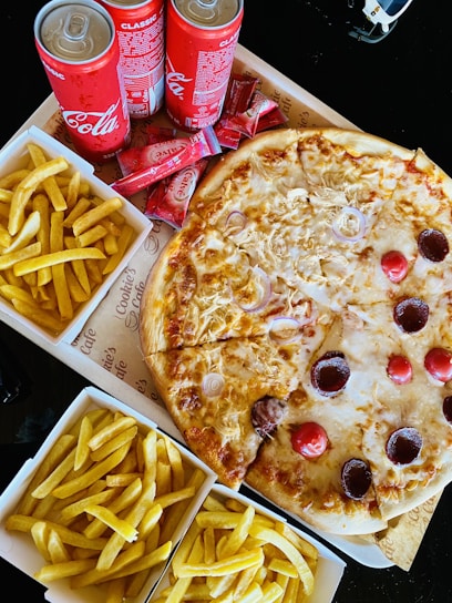 A meal spread featuring a large pizza topped with cheese, onions, and various meats. Surrounding the pizza are several boxes of golden, crispy French fries. Three cans of Coca-Cola Classic are placed nearby, along with a stack of ketchup sachets.
