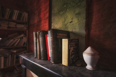 A collection of vintage books is arranged on an old, dust-covered shelf. The setting features a distressed green wall and textured red backdrop, adding an antique charm to the scene. Shelved books fill the background, while a small, ornate white urn sits nearby.