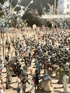 A miniature diorama depicting a historical or cultural scene with a large group of small figurines. Many figures, some wearing hats, are gathered outdoors, possibly in a market or public gathering. Sparse trees are visible, and the background is slightly blurred, giving depth to the scene.