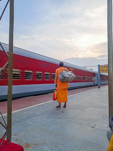 A person wearing an orange robe stands on a railway platform, carrying a large gray bag over their shoulder. The person is also holding a small red bag. A red train is passing by in the background with a sign reading 'RAJN' visible on the platform. The sky is partly cloudy, casting a soft light over the scene.