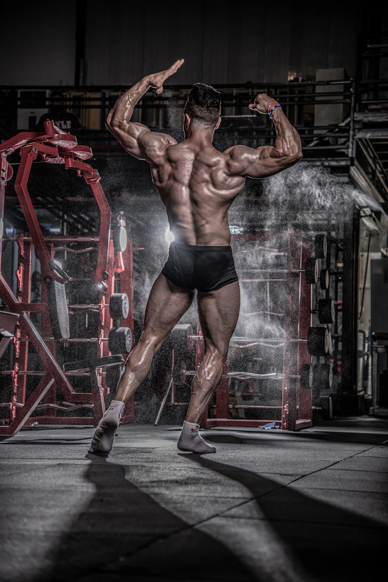 Bodybuilder posiert von hinten in einer dunklen Trainingshalle, Magnesiastaub liegt im Gegenlicht