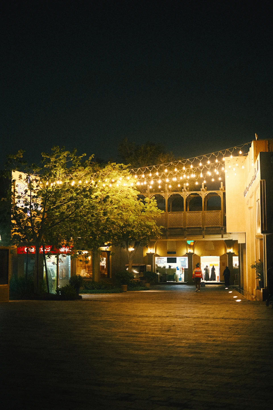 The image depicts a lively street scene at night, with a large building featuring arched windows and a balcony on its second floor adorned with string lights that cast a warm glow over the area. The building is situated between two other structures, creating an urban landscape. A solitary tree stands in front of the building, adding to the charm of the scene. Two individuals can be seen walking along the sidewalk, one carrying a bag and the other holding a camera.