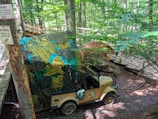 A dinosaur sculpture is positioned in a wooded area near a wooden boardwalk. Camouflage netting partially covers the dinosaur and a jeep beneath it, which is marked with the logo of a Dino Park. Signs attached to a tree provide warnings in different languages. The scene is set in a dense forest with sunlight filtering through the canopy.