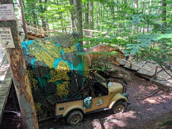 A dinosaur sculpture is positioned in a wooded area near a wooden boardwalk. Camouflage netting partially covers the dinosaur and a jeep beneath it, which is marked with the logo of a Dino Park. Signs attached to a tree provide warnings in different languages. The scene is set in a dense forest with sunlight filtering through the canopy.