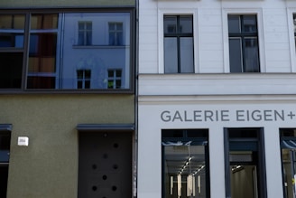 Two buildings are adjacent to each other, with different architectural styles. One building has a green facade with large windows featuring reflections of another building, while the other is white with the name 'GALERIE EIGEN+' displayed prominently above its entrance. The entrance of this gallery is characterized by modern glass doors.