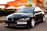 A sleek black car with white rims is parked on a street. The car has a sporty appearance, featuring a distinct front grill and headlights. In the background, there is a building with signage in Cyrillic, outdoor umbrellas, and greenery.