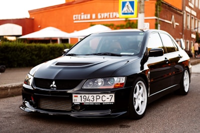 A sleek black car with white rims is parked on a street. The car has a sporty appearance, featuring a distinct front grill and headlights. In the background, there is a building with signage in Cyrillic, outdoor umbrellas, and greenery.