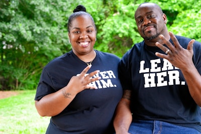 A smiling man and woman are posing together outdoors with a lush green background. Both are wearing black t-shirts with bold white text, featuring messages likely intended to be empowering or inspirational. They are making gestures with their hands, perhaps indicating numbers or a shared sign.