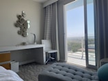 A modern hotel room includes a desk with a lamp and telephone, a decorative wall piece made of small circular mirrors, and a sleek white chair. The room features floor-to-ceiling glass doors opening onto a balcony with a view of the city and greenery. Curtains frame the doors, and the decor is minimalist yet stylish.
