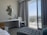 A modern hotel room includes a desk with a lamp and telephone, a decorative wall piece made of small circular mirrors, and a sleek white chair. The room features floor-to-ceiling glass doors opening onto a balcony with a view of the city and greenery. Curtains frame the doors, and the decor is minimalist yet stylish.