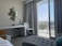 A modern hotel room includes a desk with a lamp and telephone, a decorative wall piece made of small circular mirrors, and a sleek white chair. The room features floor-to-ceiling glass doors opening onto a balcony with a view of the city and greenery. Curtains frame the doors, and the decor is minimalist yet stylish.