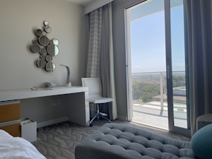 A modern hotel room includes a desk with a lamp and telephone, a decorative wall piece made of small circular mirrors, and a sleek white chair. The room features floor-to-ceiling glass doors opening onto a balcony with a view of the city and greenery. Curtains frame the doors, and the decor is minimalist yet stylish.