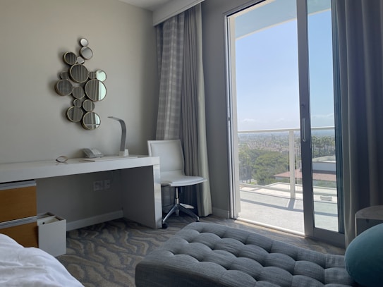 A modern hotel room includes a desk with a lamp and telephone, a decorative wall piece made of small circular mirrors, and a sleek white chair. The room features floor-to-ceiling glass doors opening onto a balcony with a view of the city and greenery. Curtains frame the doors, and the decor is minimalist yet stylish.