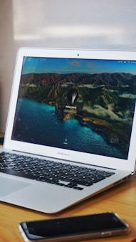 A MacBook Air is placed on a wooden desk. The laptop screen displays a picturesque wallpaper featuring a coastal landscape with rugged hills and a body of water. In front of the laptop, there is a smartphone. The overall setting appears to be a workspace.