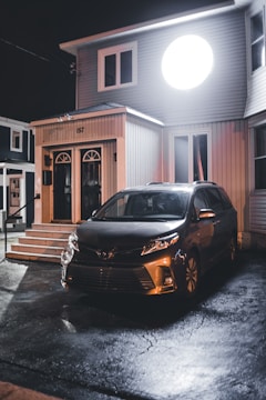 A dark, wet driveway in front of a two-story house with a large round light illuminating the facade. A car is parked on the driveway, reflecting the exterior lighting. The house has a modern design with vertical siding and arched windows near the entrance.