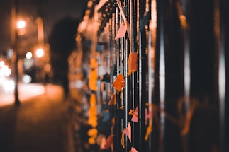 A row of colorful paper notes tied to a metal fence in a dimly lit street, with blurred lights from city traffic in the background creating a warm, soft glow.
