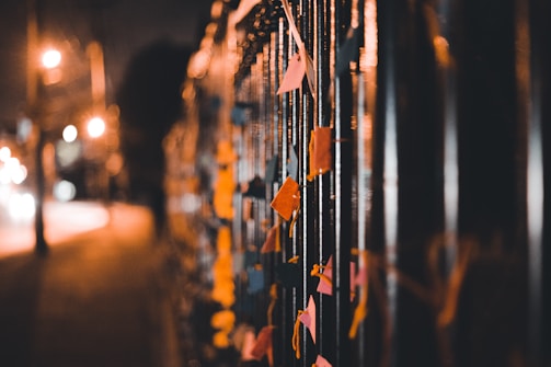 A row of colorful paper notes tied to a metal fence in a dimly lit street, with blurred lights from city traffic in the background creating a warm, soft glow.