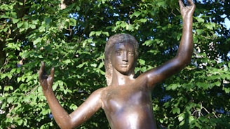 A bronze statue of a young person with raised arms set against a backdrop of lush green foliage. The statue appears to be skillfully crafted with detailed facial features and smooth surfaces, capturing a sense of grace and movement.