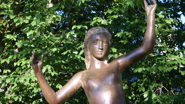 A bronze statue of a young person with raised arms set against a backdrop of lush green foliage. The statue appears to be skillfully crafted with detailed facial features and smooth surfaces, capturing a sense of grace and movement.