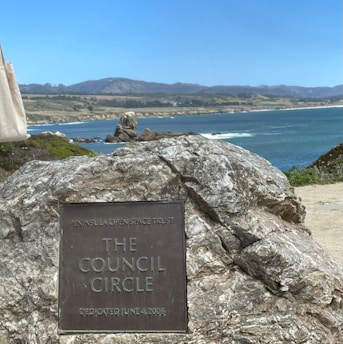 A large stone with a plaque reading 'The Council Circle' and 'DEDICATED JUNE 4, 2006' is set against a backdrop of a scenic coastal landscape. The ocean waves gently hit the rocky shoreline, with expansive hills and clear skies in the background.