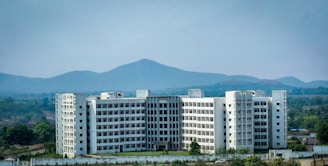 A large, multi-story white building stands prominently in the foreground, surrounded by a fence with green vegetation. The backdrop features rolling hills and a clear blue sky, creating a serene and spacious atmosphere.