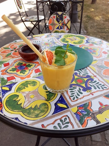 A refreshing yellow smoothie served in a glass with a straw, garnished with mint leaves and a slice of grapefruit. It is placed on a vibrant, colorful mosaic table with intricate artistic patterns. A rustic wooden ashtray and a folded chair can be seen in the background, adding to the outdoor, casual café setting.