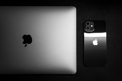 A top view of a closed silver laptop and a black smartphone placed side by side on a dark surface, both featuring a prominent Apple logo.