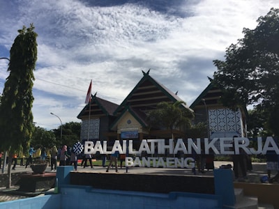 A traditional building with a peaked roof is surrounded by trees and a group of people. The structure includes patterns on the walls, and there is a large sign reading 'Balaiklatihan Kerja Bantaeng' in front.