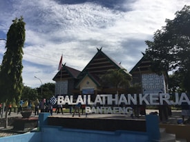 A traditional building with a peaked roof is surrounded by trees and a group of people. The structure includes patterns on the walls, and there is a large sign reading 'Balaiklatihan Kerja Bantaeng' in front.