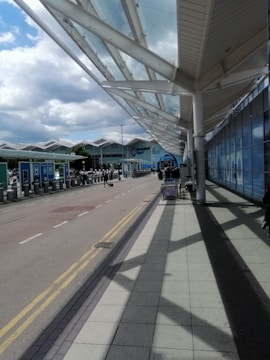 A modern, open-air airport terminal with a wide walkway covered by a glass canopy. The sky is partly cloudy, and the architecture features large glass panels and white structural supports. Luggage trolleys are visible along the walkway, and a few people are walking toward the building.