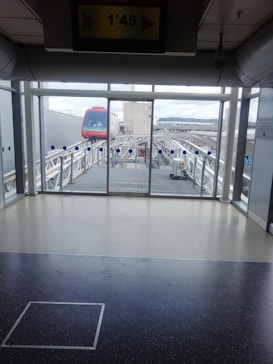 A monorail with a bright red front is visible through large glass windows on an elevated track. The indoor area features a tiled floor and some overhead signage indicating the shuttle's timing. The railing and the glass have a clean, modern design, and outside, there are cloudy skies visible.
